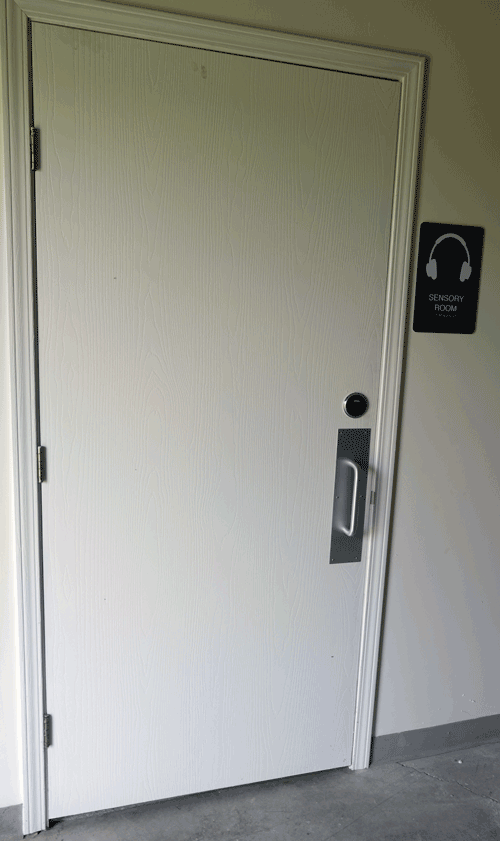 A photo of a white door with a black sign with the words "sensory room" and the symbol of noise cancelling headphones.