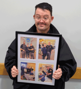 A man with dark hair and glasses holds up a picture frame with four photos in it. Two of the photos are from the Licking County Sheriff's Office Citizen's Academy graduation and the other two are deputies visiting him in the hospital.