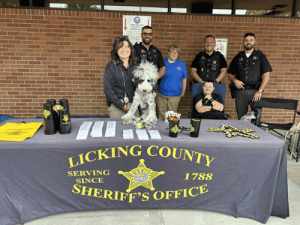 A group of sheriff's deputies and a gray and white therapy dog stand around a man who is seated in a chair. They are behind a table with the Licking County Sheriff's Office table cloth and giveaway items.