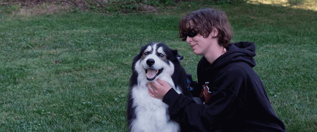 A young woman in a black hoodie kneel downs to pet her dog. The dog is a black and white dog with lots of fur. They are outside in the grass with a tree in the background.