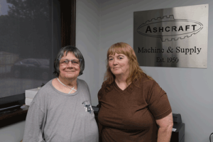 A woman in a gray shirt stands next to a woman in a brown shirt in front of an Ashcraft sign