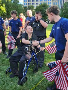 A photo of a man seated in a blue walker, shaking hands with a young man holding American flags. The deputy behind him is moved to tears and wiping his eyes.