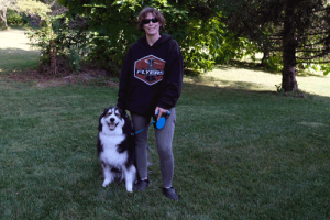 A young woman in a black hoodie stands next to her dog. The dog is a black and white dog with lots of fur. They are outside in the grass with a tree in the background.