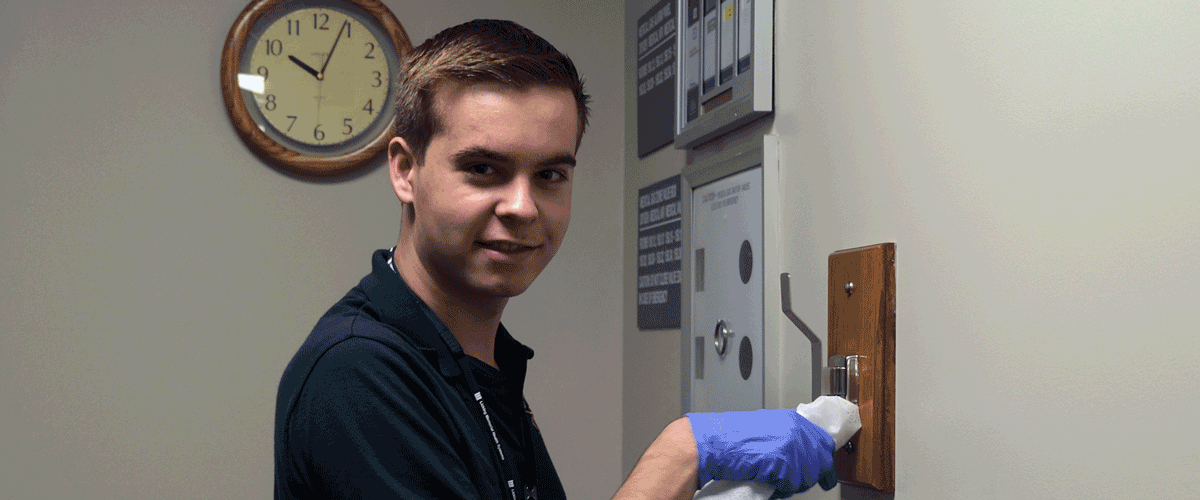 A young man in a green shirt wearing gloves uses a cloth to wipe down a hanger.