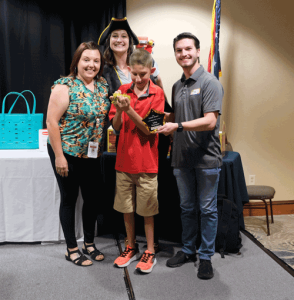 A picture of a woman in a flowered shirt, a woman in a pirate costume standing with a boy in a red polo shirt and a man in a gray polo shirt holding a star award plaque.