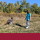 A photo of a woman walking in a field holding the leash of a gray and white fluffy dog.