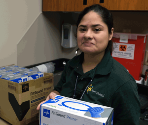 A young woman wearing a Project SEARCH polo shirt holds a box of gloves and looks at the camera.