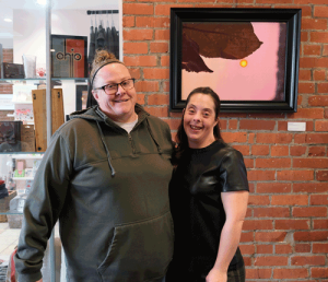 Alexis, who is on the right, stands in front of one of her photographs with her arm around her service coordinator, who is wearing a green sweatshirt and glasses.