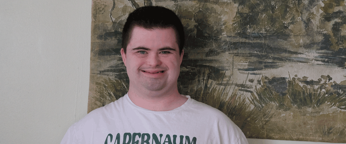 A photo of a young man with short dark hair standing in front of a green wall hanging.