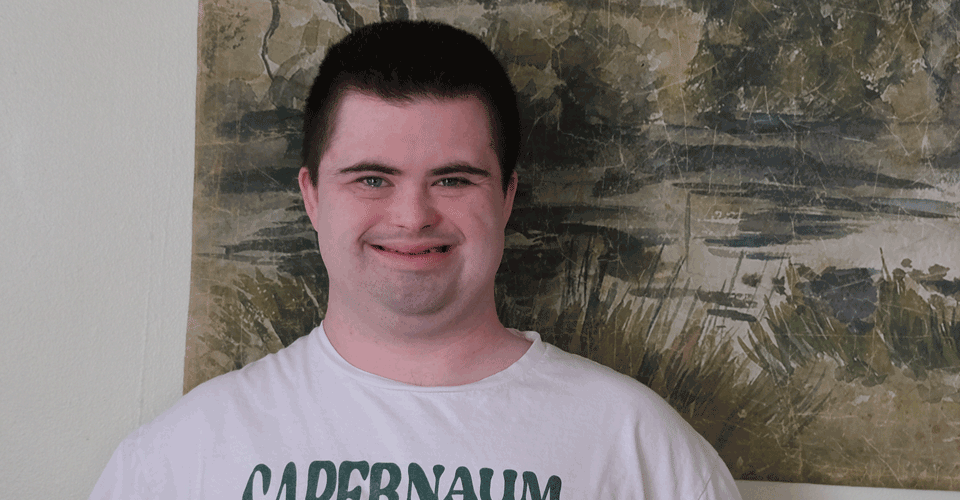 A photo of a young man with short dark hair standing in front of a green wall hanging.
