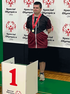 A young man with a medal around his neck stands in front of the first place podium at a Special Olympics Ohio event.
