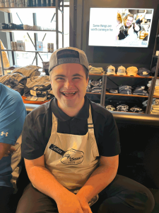 A photo of a young man in a backwards baseball cap wearing a Bitty and Beau's apron. He has a huge smile on his face and is sitting in a coffee house.