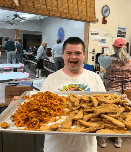 A photo of a young man with a huge smile on his face, holding a giant tray of curly friends and chicken fingers in a cafeteria.