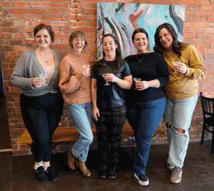 Four Hey Babes employees stand beside Alexis Ackerley at her artshow opening. They are holding glasses to toast her. They are all smiling.