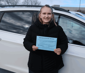 A close up image of a woman wearing a black coat stands in front of her car holding a blue envelope.