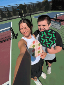 A photo of a young man holding a green pickleball paddle on a pickleball court. His sister is standing in front of him, holding her own paddle and taking a selfie.