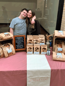 A young man with a "Bread Boy" shirt has his arm around his sister.They are standing behind a table of sourdough bread and cookies. There is a pink and white Sundae Loaf tablecloth on the table.