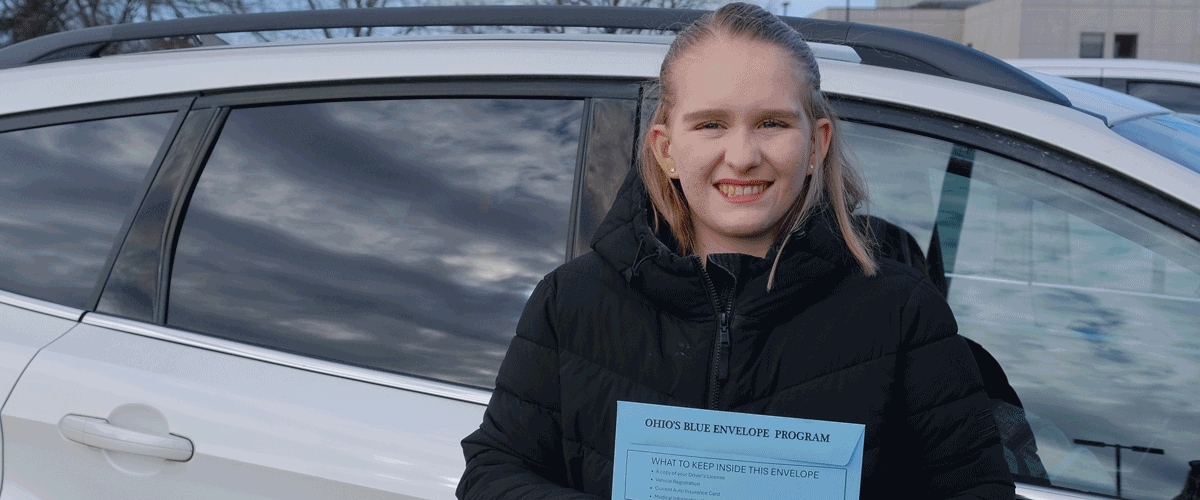 A woman wearing a black coat stands in front of her car holding a blue envelope.