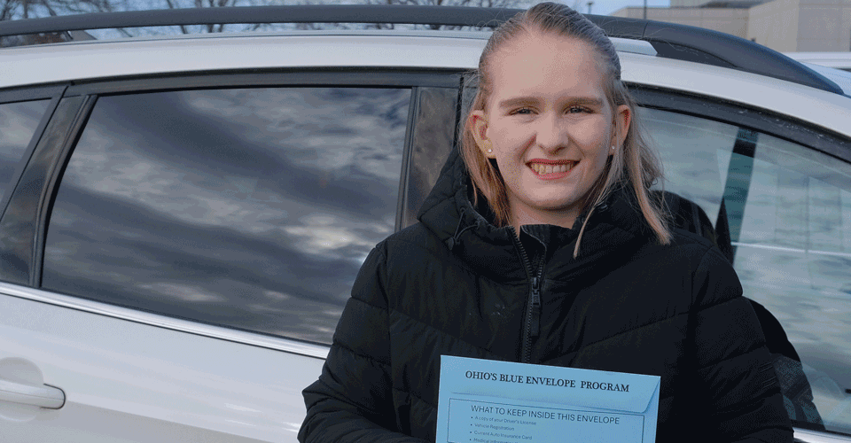 A woman wearing a black coat stands in front of her car holding a blue envelope.