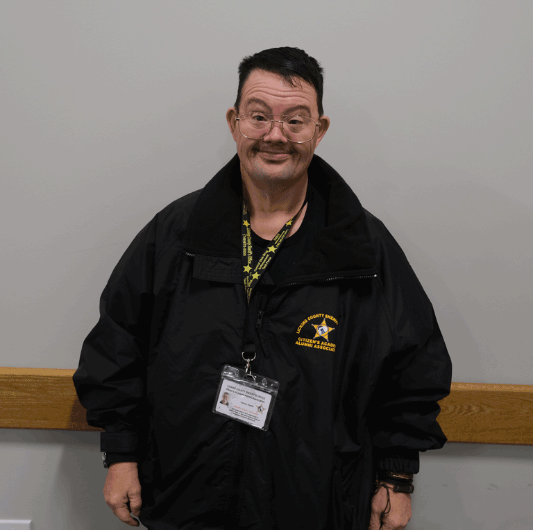 A man with dark hair and glasses smiles at the camera. He is wearing a black jacket and a Licking County Sheriff's Office lanyard.