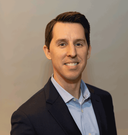 A headshot of Alex Corwin. He is a white man with dark hair wearing a light blue shirt and a dark colored blazer.