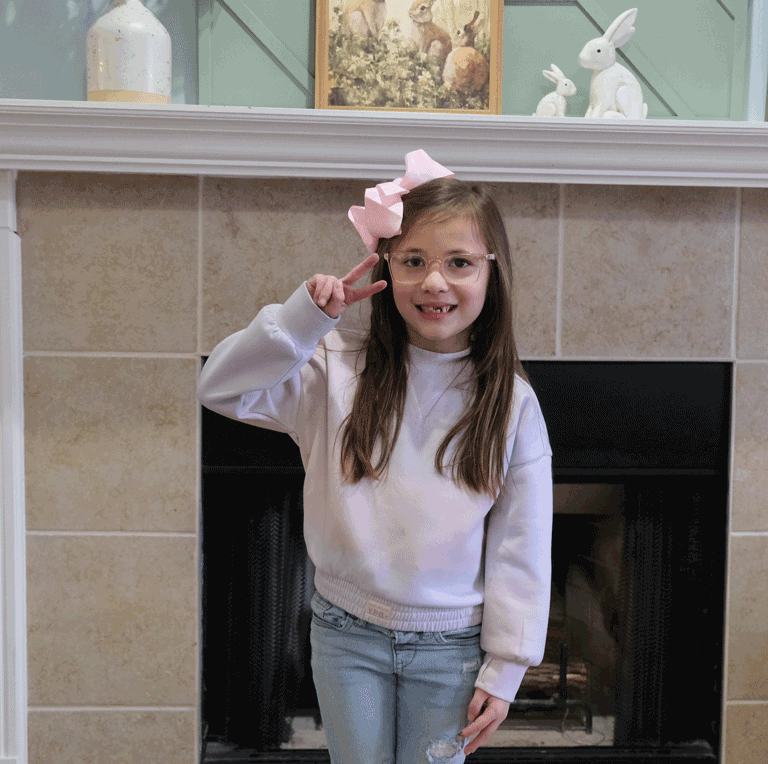A little girl with long hair stands in front of her fireplace at home. She has on a pink sweater and a pink hair bow. She is holding up her fingers in a peace sign.