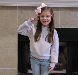 A little girl with long hair stands in front of her fireplace at home. She has on a pink sweater and a pink hair bow. She is holding up her fingers in a peace sign.