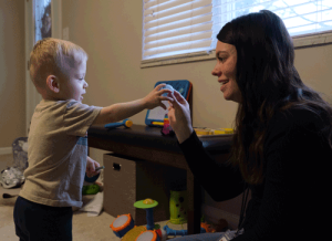 A little boy with blonde hair holds a pingpong ball out and a woman with long dark hair takes it from him.