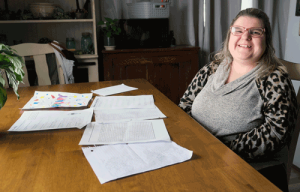 A woman in a leopard print shirt who is wearing classes sits at a table, showing off completed worksheets. 