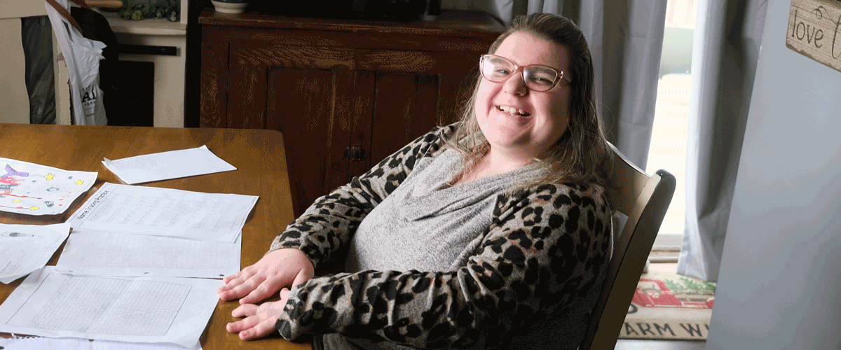 A woman in a leopard print shirt who is wearing classes sits at a table, showing off completed worksheets. She has a big smile on her face.