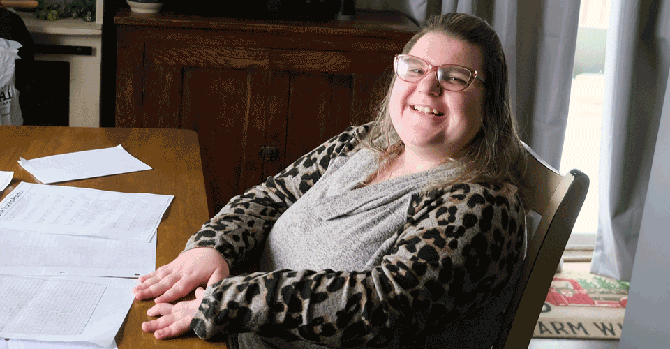 A woman in a leopard print shirt who is wearing classes sits at a table, showing off completed worksheets. She has a big smile on her face.