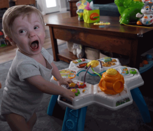 A little boy in a onesie uses a standing toy to stand up. He has his mouth open in a silly face.