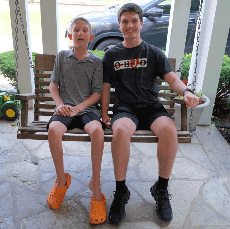A photo of a young boy sitting on a porch swing, next to a 19-year old man wearing an OSU shirt. They are smiling and looking at the camera.
