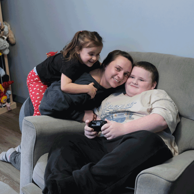 A boy sits in a chair and gets a hug from his mother and little sister.