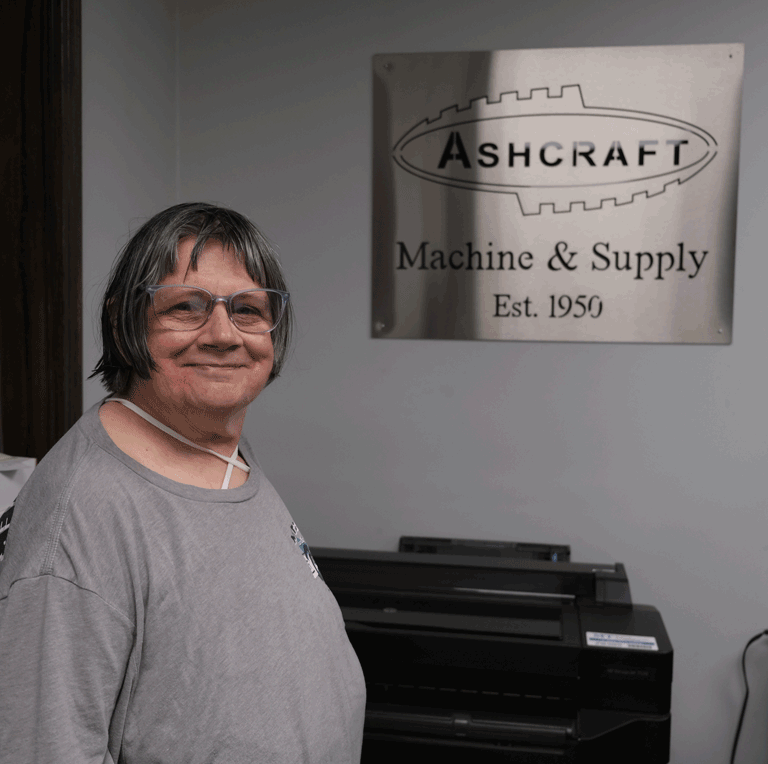 A woman with glasses and a gray shirt stands in front of a metal Ashcraft sign.