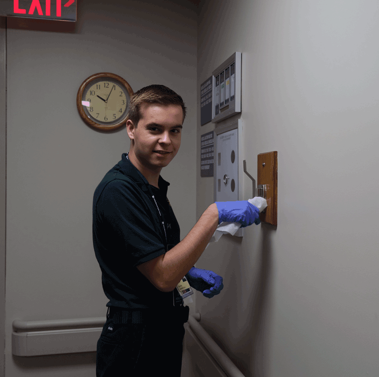 A young man in a green shirt wearing gloves uses a cloth to wipe down a hanger.