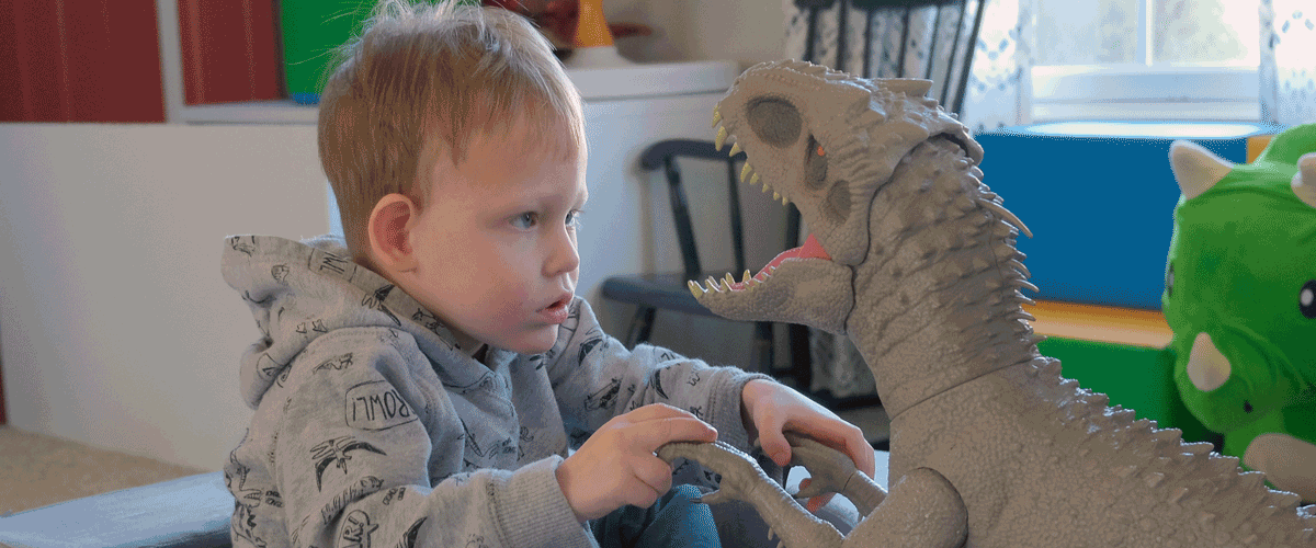 A little boy sits and looks thoughtfully at a large toy dinosaur.