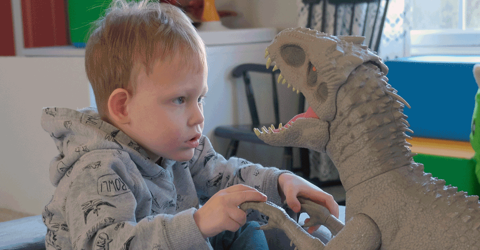 A little boy sits and looks thoughtfully at a large toy dinosaur.