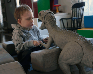 A little boy sits and looks thoughtfully at a large toy dinosaur. He is holding the dinosaurs hands in his.