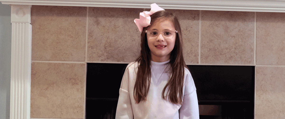 A little girl with long hair stands in front of her fireplace at home. She has on a pink sweater and a pink hair bow.