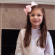 A little girl with long hair stands in front of her fireplace at home. She has on a pink sweater and a pink hair bow.