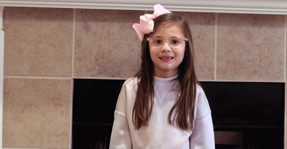 A little girl with long hair stands in front of her fireplace at home. She has on a pink sweater and a pink hair bow.