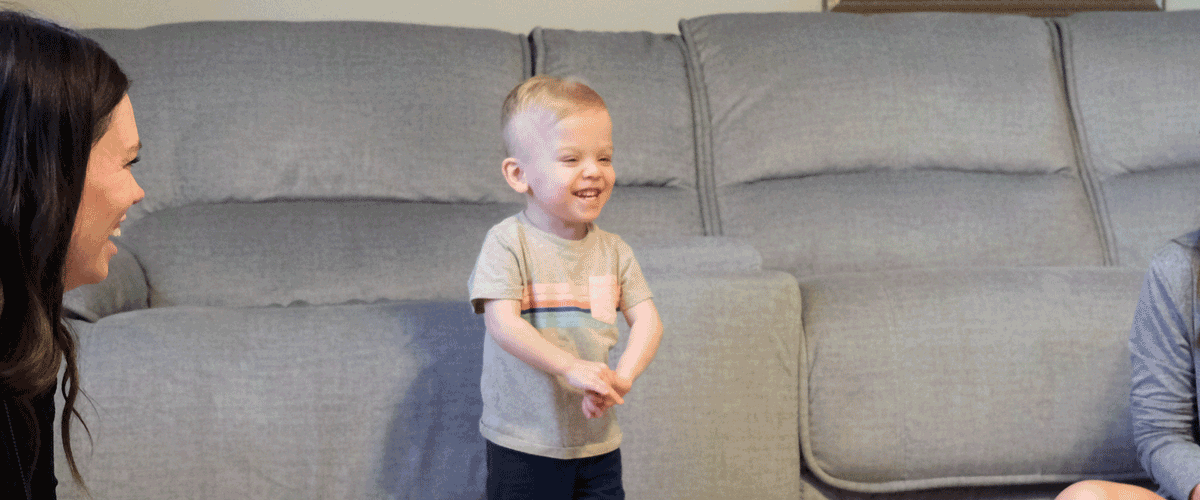 A little boy with blonde hair smiles widely. His mother and LCBDD Occupational Therapist smile at him.