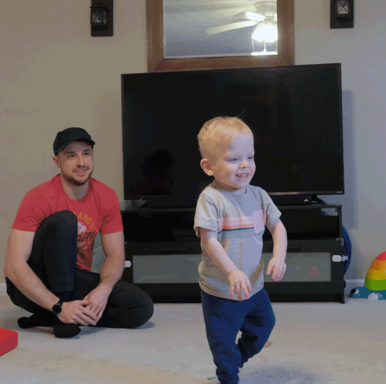 A little boy with blonde hair walks in his living room. His father is sitting behind him and smiling.
