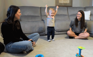 A little boy raises his arms in celebration. His mother and LCBDD Occupational Therapist smile at hime.