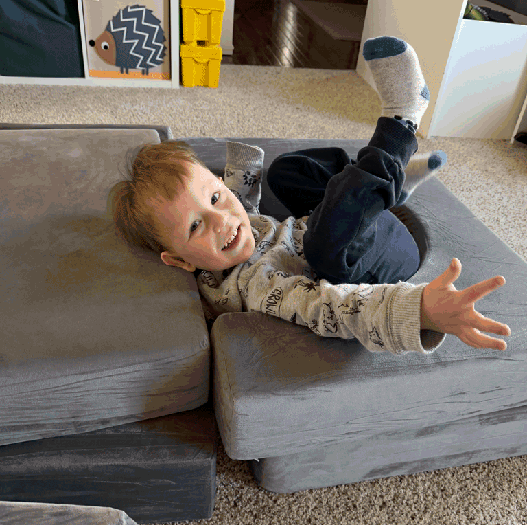 A little boy flops down on couch cushions with a smile. He has dark blonde hair and is wearing a dinosaur sweatshirt and blue pants.