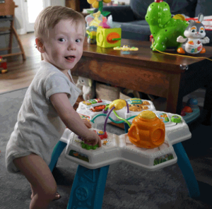 A little boy in a onesie uses a standing toy to stand up. He has a mischievous look on his face.