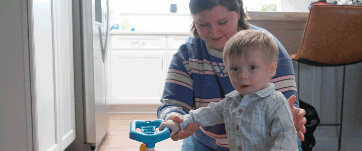 A woman in a striped shirt stands behind a toddler with blonde hair. He is holding a firetruck toy.