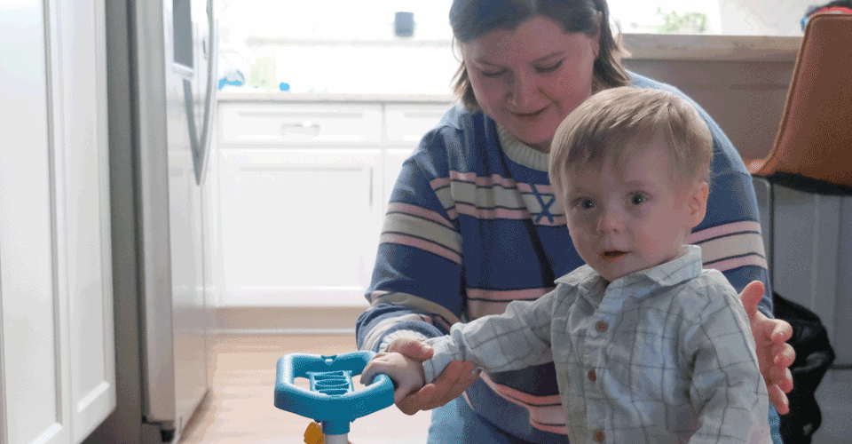 A woman in a striped shirt stands behind a toddler with blonde hair. He is holding a firetruck toy.