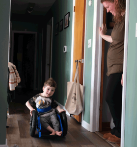 A little boy wheels his GoBro wheelchair down the hallway. His mother stands at the kitchen door smiling down at him.